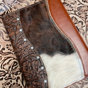 May include: A brown and white cowhide leather notebook cover with a brown leather flap and turquoise stitching. The cover has a floral embossed design and pearl-like studs.