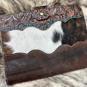 May include: A brown leather planner with a cowhide design and turquoise stitching. The planner has a flap closure with a brass button and the word "Junaco" is embossed on the front.