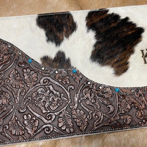 May include: A brown leather placemat with a floral embossed design. The placemat has a white and brown cowhide patch with the initials "KR" embroidered on it.