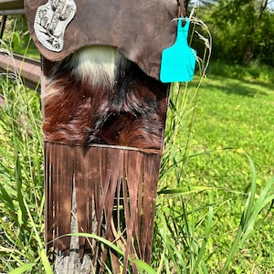 May include: A brown leather crossbody bag with a fringe bottom. The bag has a white and brown cowhide flap with a cactus embroidery. The bag is hanging on a wooden post.
