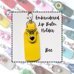 May include: A yellow embroidered lip balm holder featuring a bee design and a black heart. It has a silver clasp and the text "Embroidered Lip Balm Holder" and "Bee". Other holders with various designs are in the background.