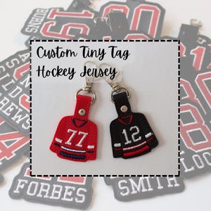 Peut inclure: Deux porte-clés miniatures en forme de maillot de hockey, l'un rouge avec le numéro 77 et l'autre noir avec le numéro 12. Les porte-clés sont attachés à des fermoirs argentés. Le texte sur l'image indique "Custom Tiny Tag Hockey Jersey."