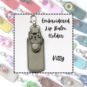 Könnte beinhalten: Ein grauer, bestickter Lippenbalsamhalter mit einem Katzengesicht-Design. Der Halter hat einen Druckknopfverschluss und einen silberfarbenen Clip. Der Text "Embroidered Lip Balm Holder" und "Kitty" ist abgebildet.