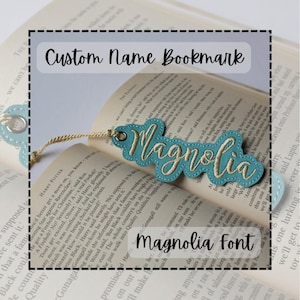 Puede incluir: Marcapáginas azul claro con el nombre "Magnolia" bordado en una fuente cursiva. El marcapáginas tiene un borde festoneado y un cordón beige. También se ve el texto "Custom Name Bookmark" y "Magnolia Font".