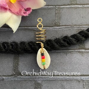 May include: A gold-toned metal hair coil accessory featuring a white cowrie shell charm accented with small black, red, yellow, and green beads. The hair jewelry is wrapped around a thick black braided hair strand. Text reads OrchidWay Treasures.
