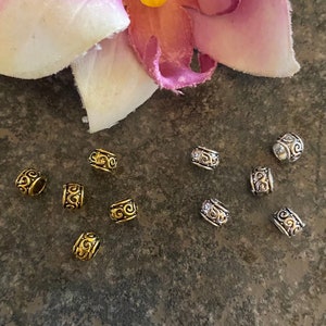 May include: A collection of gold and silver metal beads with a decorative swirl design. The beads are arranged on a textured surface.