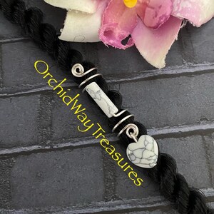 May include: Black hair braid adorned with silver wire wraps, a white and gray stone bead, and a heart-shaped charm. The text "Orchidway Treasures" is visible in yellow.