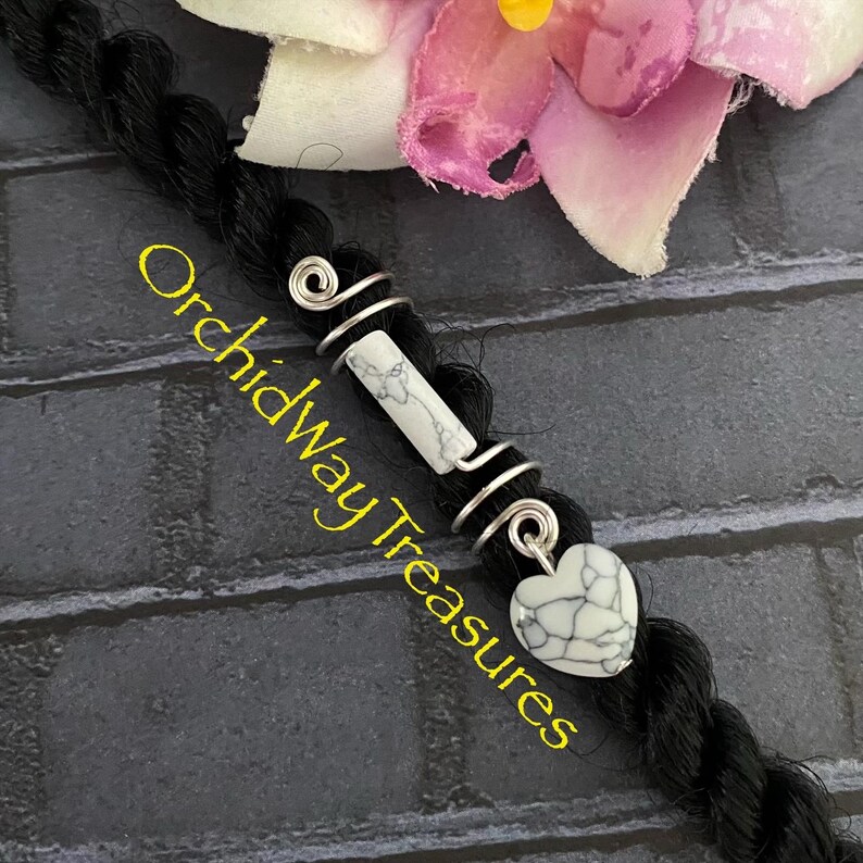 May include: Black hair braid adorned with silver wire wraps, a white and gray stone bead, and a heart-shaped charm. The text "Orchidway Treasures" is visible in yellow.