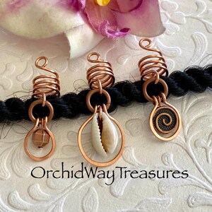 May include: Three copper hair jewelry pieces with spiral and teardrop designs, one with a shell, one with a bead, and one with a spiral. The jewelry is attached to a black braid. The text "OrchidWay Treasures" is at the bottom.