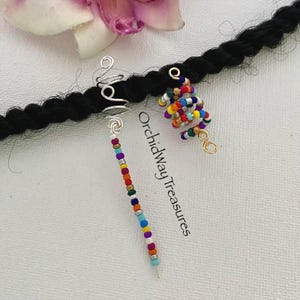 May include: Close-up of three hair accessories: a silver spiral, a beaded strand, and a spiral of colorful beads. The accessories are on a black braid, with a white background and a pink flower. The text "Orchidway Treasures" is visible.