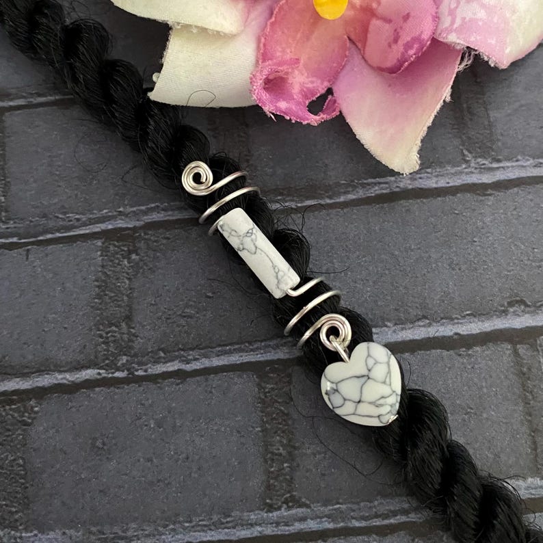 May include: A black hair braid adorned with silver wire wraps and white stone accents. The design includes a rectangular stone and a heart-shaped charm, creating a unique hair accessory. A pink and white flower is in the background.