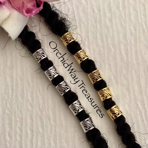 May include: Two strands of black hair with silver and gold metal beads. The beads have a decorative, swirling pattern. The text "Orchidway Treasures" is visible on the image.