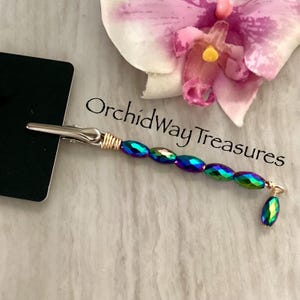 May include: A silver-toned alligator clip with a gold-colored wire wrap and a strand of iridescent, oval-shaped beads. A single, matching bead dangles from the end. The text "OrchidWay Treasures" is visible.