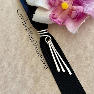 May include: A silver charm with three dangling strands of silver wire. The charm is attached to a black ribbon with the text "Orchidway Treasures" printed on it.