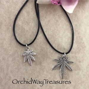May include: Two necklaces with black cords and silver-colored marijuana leaf pendants. The pendants have detailed veining. The text "OrchidWay Treasures" is at the bottom of the image.
