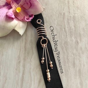 May include: A black ribbon hair accessory with a copper-colored wire spiral and dangling beads. The accessory is adorned with a pink and purple orchid flower. The text "OrchidwayTreasures" is printed on the ribbon.