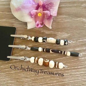 May include: Three beaded hair clips with silver-colored metal clips. The beads are in various colors, including black, white, and brown. The text "OrchidWayTreasures" is visible at the bottom of the image. A pink and purple orchid flower is in the background.