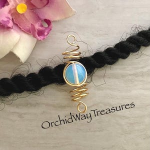 May include: A gold-colored hair accessory with a spiral design, featuring a round, light blue and white stone. The accessory is placed on a black braid, with the text "OrchidWay Treasures" in the bottom right corner.