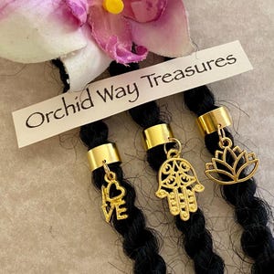 May include: Close-up of black hair strands adorned with gold-colored hair cuffs and charms. Charms include a heart with the word "LOVE", a Hamsa hand, and a lotus flower. The text "Orchid Way Treasures" is visible.