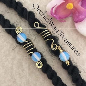 May include: Two gold wire hair beads with opal beads. The beads are on black dreadlocks. The text "Orchidway Treasures" is visible in the background.