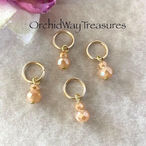 May include: Four gold stitch markers with peach-colored glass beads. The beads are faceted and have a pearlescent finish. The stitch markers are a popular crafting supply.