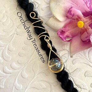 May include: A gold-colored wire hair wrap with a teardrop-shaped pendant holding a clear, round bead. The wrap is on a black braid, with the text "Orchidway Treasures" visible. A pink flower is in the background.