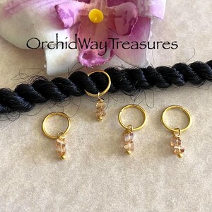 May include: Four gold hair tie beads with a clear, light brown crystal charm. The beads are shown on a black braid.