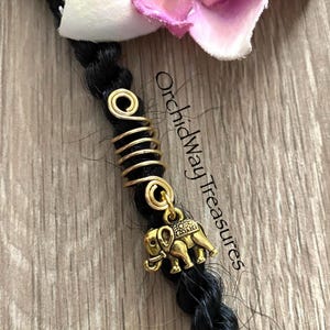 May include: Gold hair beads with a small elephant charm. The beads are coiled and the charm is attached to the bottom of the beads. The beads are on a black braid. The text "OrchidWay Treasures" is visible in the background.