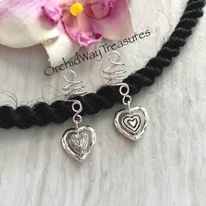 May include: Two silver heart-shaped hair charms with a spiral design, attached to a black braid. The charms have a textured heart within a heart design. A pink and yellow flower is in the background. The text "OrchidWay Treasures" is visible.