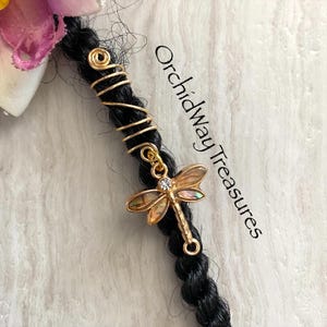 May include: A gold-colored dragonfly charm with iridescent wings and a small crystal accent, attached to a black braid with gold wire wraps. The text "Orchidway Treasures" is visible.