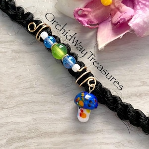 May include: A black braid adorned with gold wire wraps, colorful beads, and a mushroom charm. The charm is blue with yellow, orange, and white spots. The text "Orchidway Treasures" is visible.