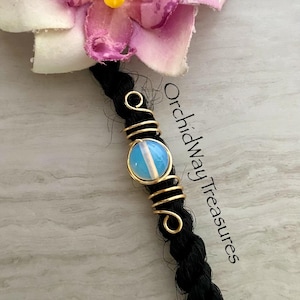 May include: A gold-toned wire hair wrap with a light blue opal-like bead, wrapped around a black braid. The text "Orchidway Treasures" is visible. A pink and white flower is in the background.