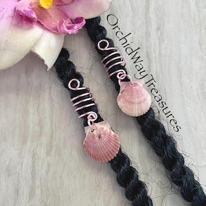 May include: Two hair accessories featuring rose gold wire coils wrapped around black braided hair strands. Each coil is attached to a small, ribbed, light pink seashell charm. The text OrchidWayTreasures is visible in black script.