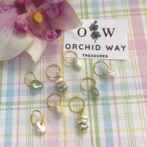 May include: Nine gold stitch markers with different colored pearl beads. The markers are on a pink and green checkered background. The text "Orchid Way Treasures" is on a white card.