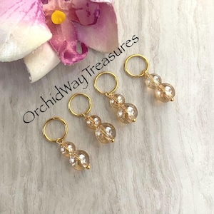 May include: Four gold-colored stitch markers with clear, amber-toned beads. Each marker has a small gold ring at the top. The background includes a pink orchid and the text "OrchidWay Treasures".