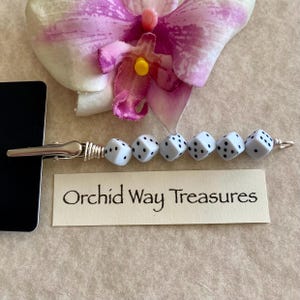 May include: A silver clip with a chain attached to a string of white dice beads with black dots. The clip is attached to a card that reads "Orchid Way Treasures".