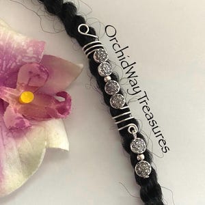 May include: Silver dreadlock beads with a decorative design. The beads are strung on a silver wire that is wrapped around a dreadlock.