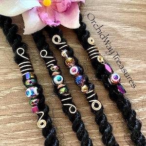 May include: Four gold hair beads with colorful beads and a gold wire wrap. The beads are iridescent and have a white eye design. The beads are on black dreadlocks. The text "OrchidWayTreasures" is visible in the image.