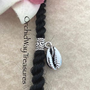 May include: Close-up of a black dreadlock with a silver-colored, ornate bead and a seashell charm. The text "Orchidway Treasures" is visible on the left side of the image. The background is a neutral, light beige color.