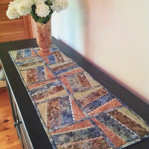 May include: A rectangular quilted table runner with a patchwork design in shades of blue, brown, and beige. The runner is placed on a dark surface, with a vase of white flowers in the background.