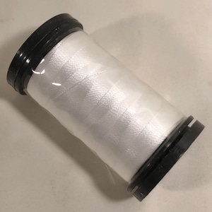 May include: A spool of white thread, tightly wound and encased in a clear plastic wrap. The spool has black plastic caps on each end. The thread appears to be a smooth, shiny material, suitable for sewing or crafting projects.