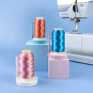 Op de afbeelding: Drie spoelen garen in oranje, blauw en roze, elk op een verschillend gekleurde houder, worden getoond voor een naaimachine. De naaimachine is wit en zilverkleurig, met de merknaam "BERNINA" zichtbaar.
