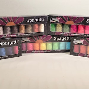 May include: Five boxes of "Spagetti" thread sets are displayed. Each box features a variety of thread colors, visible through a clear window. The boxes have a dark background with a floral design and the brand name. The thread colors range from neutral to vibrant.