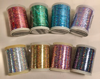 Puede incluir: Ocho carretes de hilo holográfico en varios colores, incluyendo rosa, verde, azul y plateado. Cada carrete está etiquetado con la palabra "Hologram" y "Wonderfill". El hilo está enrollado en carretes blancos con tapas de plástico transparente.