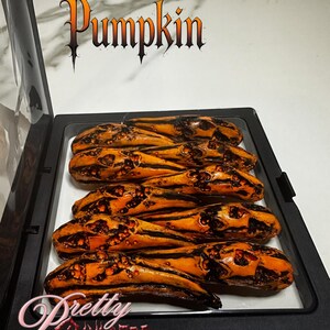Peut inclure: Un présentoir noir contenant six doigts en forme de citrouille orange et noire. Le présentoir porte les inscriptions "Pumpkin" et "Pretty Sinister".