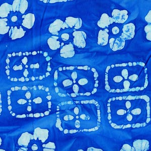 May include: A vibrant blue fabric featuring a repeating pattern of white, hand-dyed floral motifs and rectangular shapes containing four-petaled flower designs. The textile displays a distinct batik-style aesthetic with soft, irregular edges.
