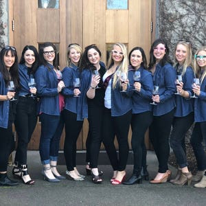 May include: A group of women wearing denim shirts and black trousers are holding wine glasses. They are standing in front of a wooden door with a stone wall behind them.