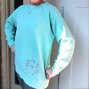 May include: A turquoise blue sweatshirt with a silver embroidered monogram "SRL" on the left side.