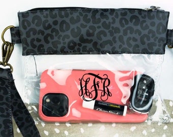 May include: A clear plastic crossbody bag with a black leopard print flap. The bag has a zipper closure and a long adjustable strap. The bag is filled with a pink phone case with a monogram, a lip balm, and a key chain.