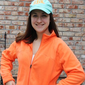 May include: An orange fleece jacket with a full zipper and side pockets. The person is wearing a light blue baseball cap with the embroidered letters "BBM". The background features a brick wall and outdoor foliage.